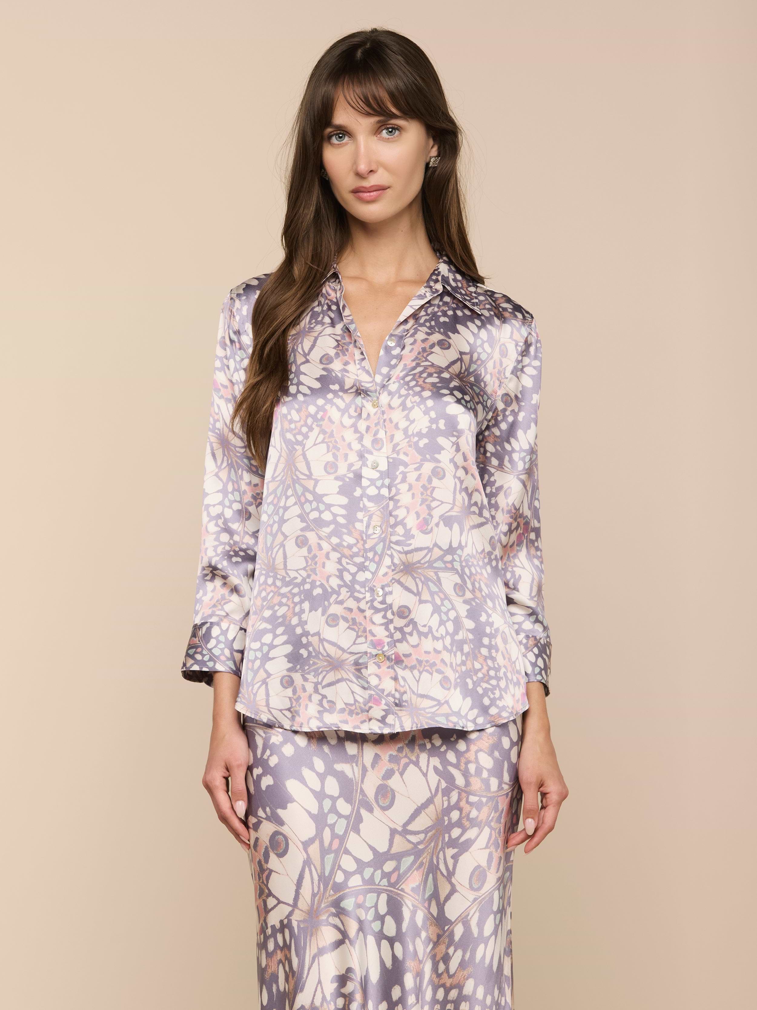 Una donna dai lunghi capelli castani modella la blusa di seta Dani e la gonna abbinata in morbide tonalità pastello con stampa di ali di farfalla, in piedi su un semplice sfondo beige e rivolta in avanti con unespressione neutra.