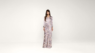 A woman stands in a long, patterned dress with long sleeves against a plain white background, looking directly at the camera.
