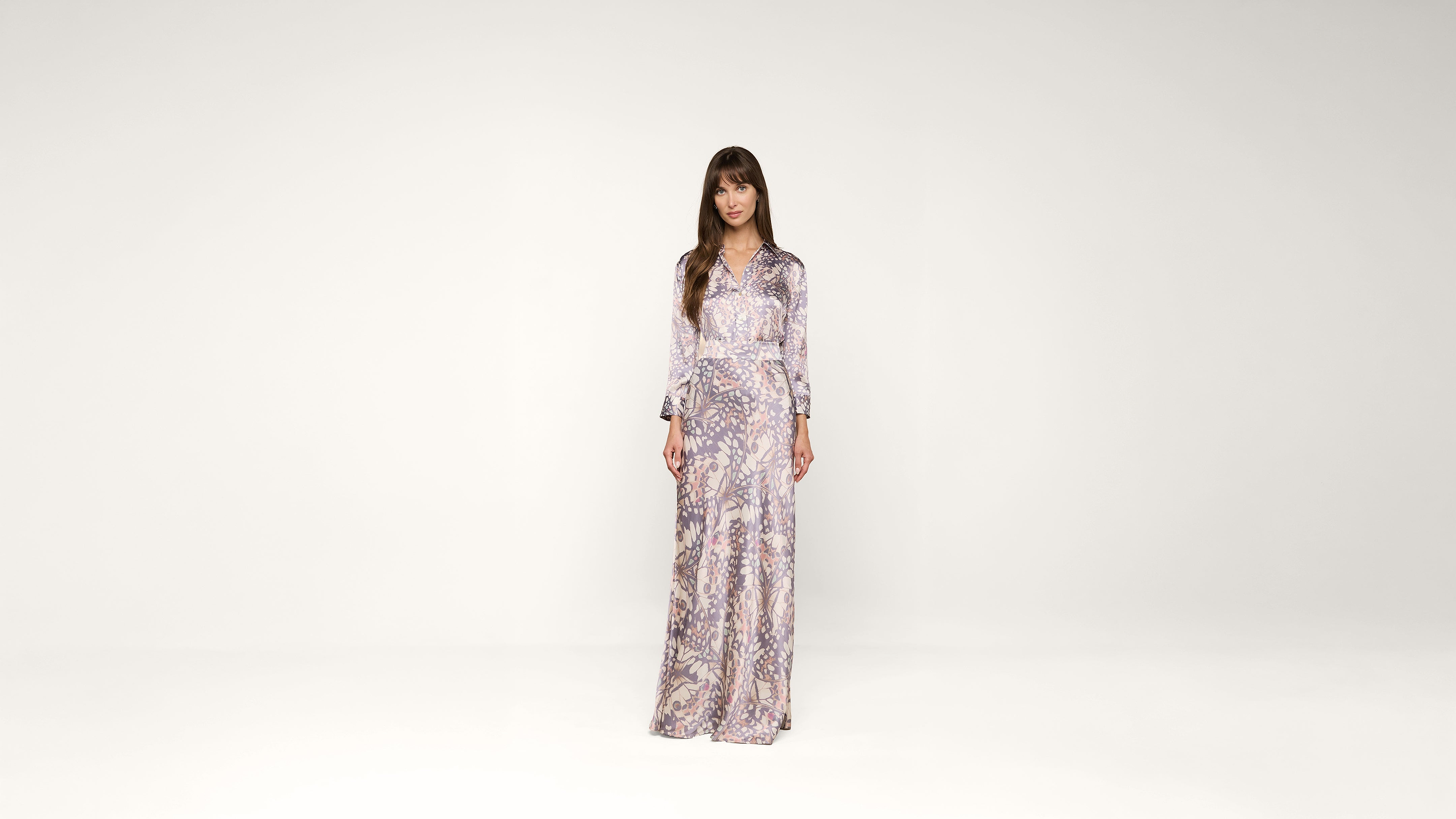 A woman stands in a long, patterned dress with long sleeves against a plain white background, looking directly at the camera.