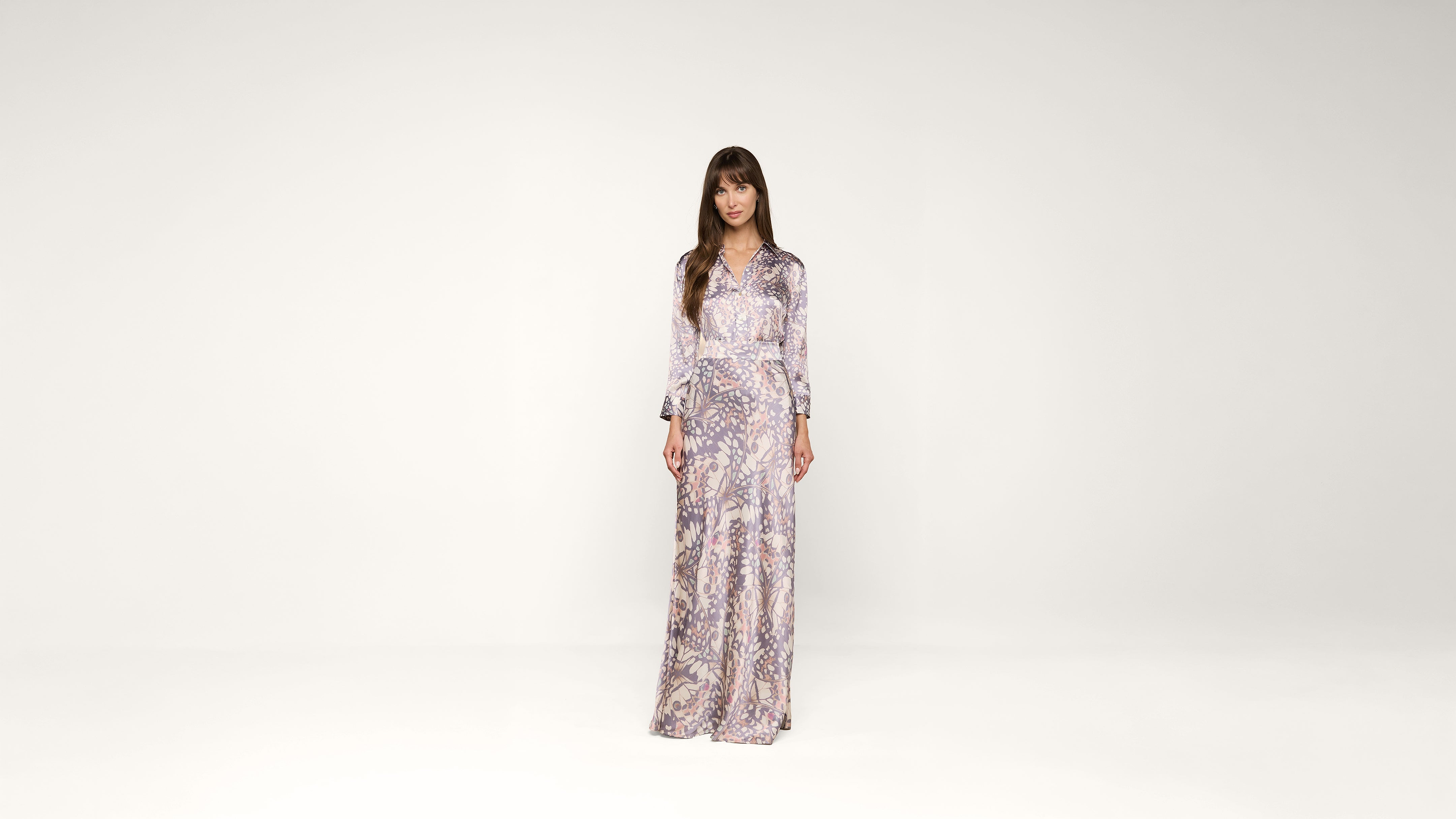A woman stands in a long, patterned dress with long sleeves against a plain white background, looking directly at the camera.