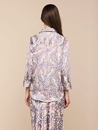 Una donna dai lunghi capelli castani è in piedi, rivolta verso lesterno, e indossa la blusa Dani Silk con stampa di ali di farfalla e maniche a tre quarti e pantaloni abbinati su uno sfondo beige.