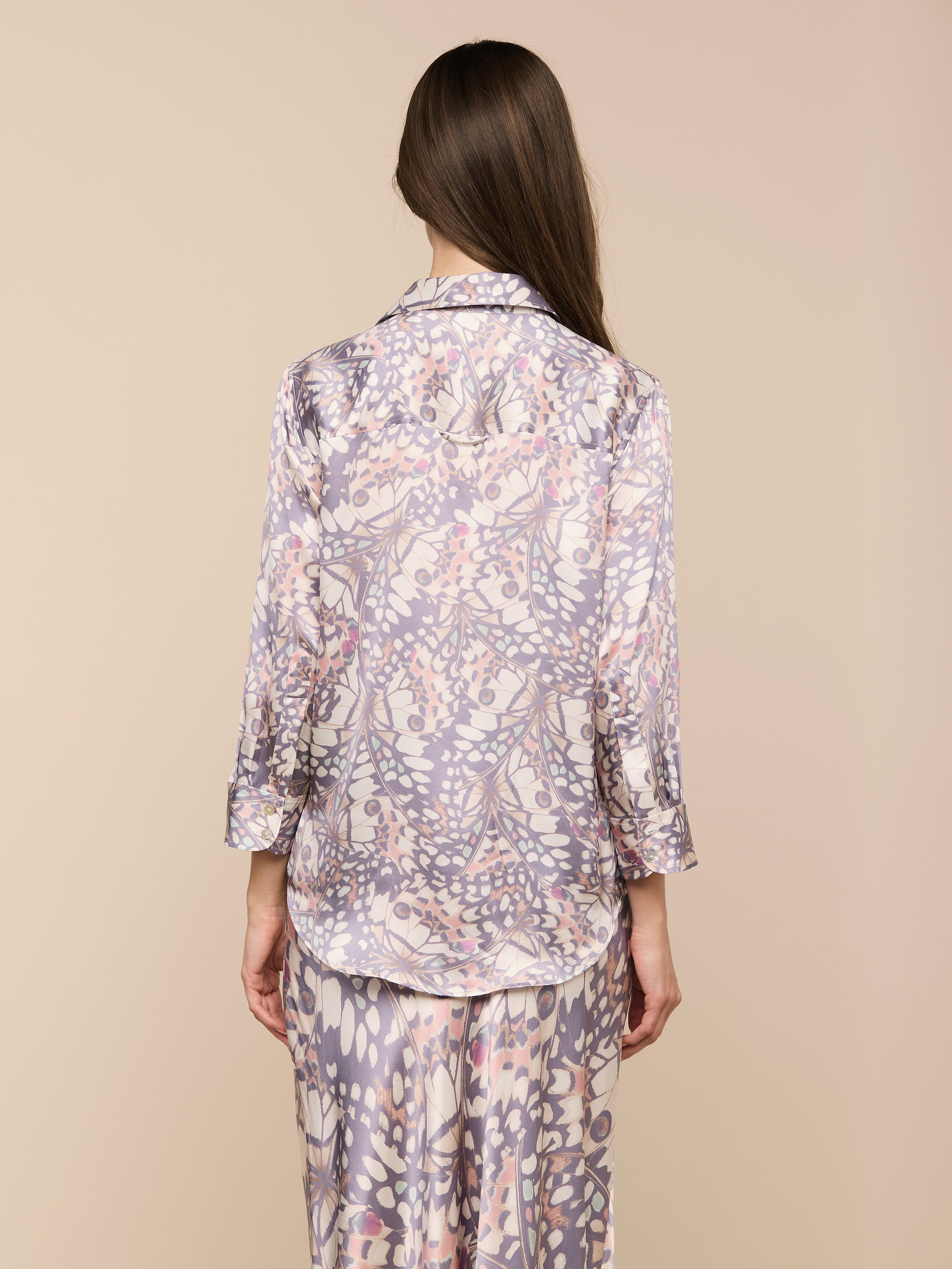 Una donna dai lunghi capelli castani è in piedi, rivolta verso lesterno, e indossa la blusa Dani Silk con stampa di ali di farfalla e maniche a tre quarti e pantaloni abbinati su uno sfondo beige.