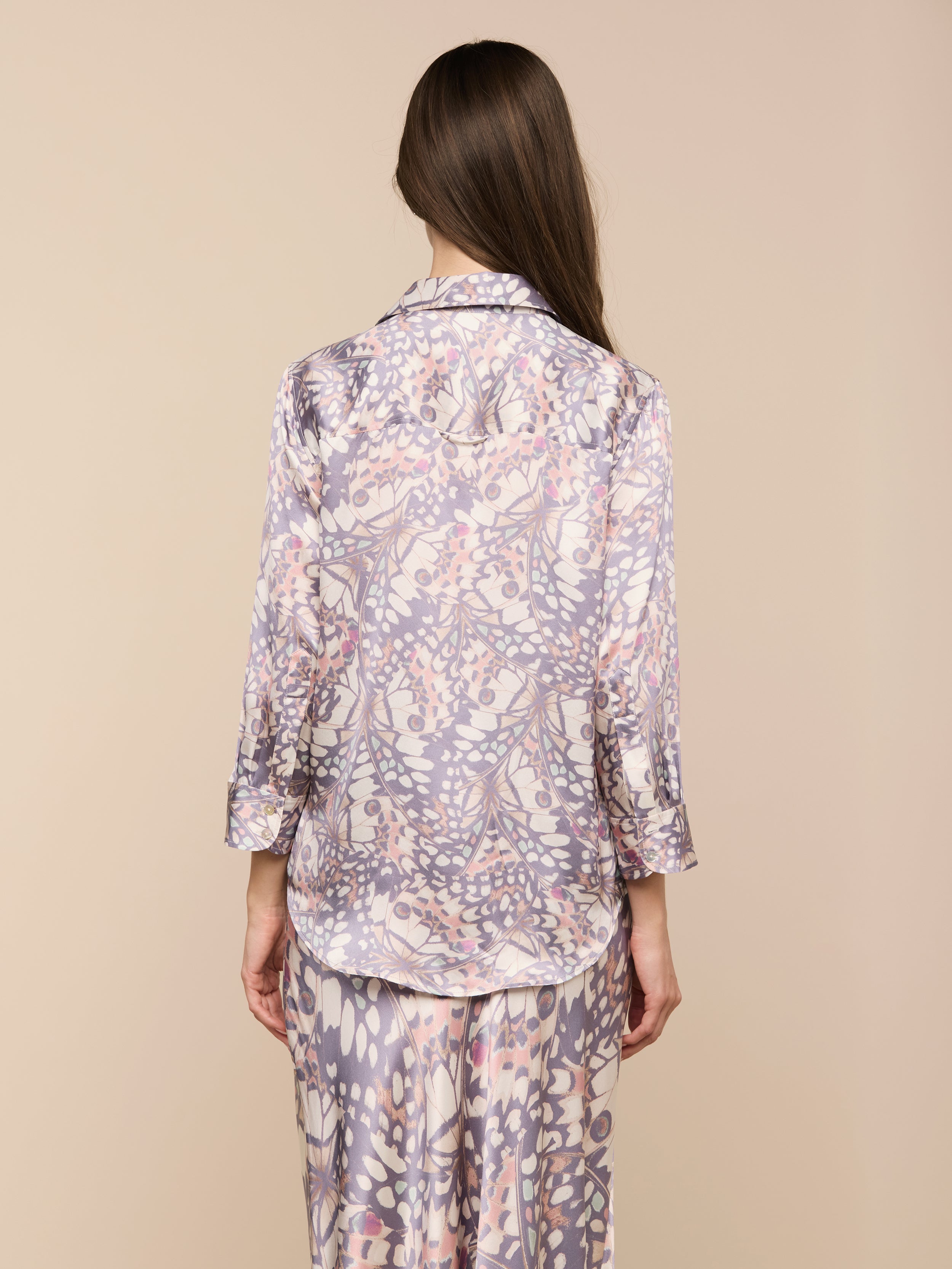 Una donna dai lunghi capelli castani è in piedi, rivolta verso lesterno, e indossa la blusa Dani Silk con stampa di ali di farfalla e maniche a tre quarti e pantaloni abbinati su uno sfondo beige.