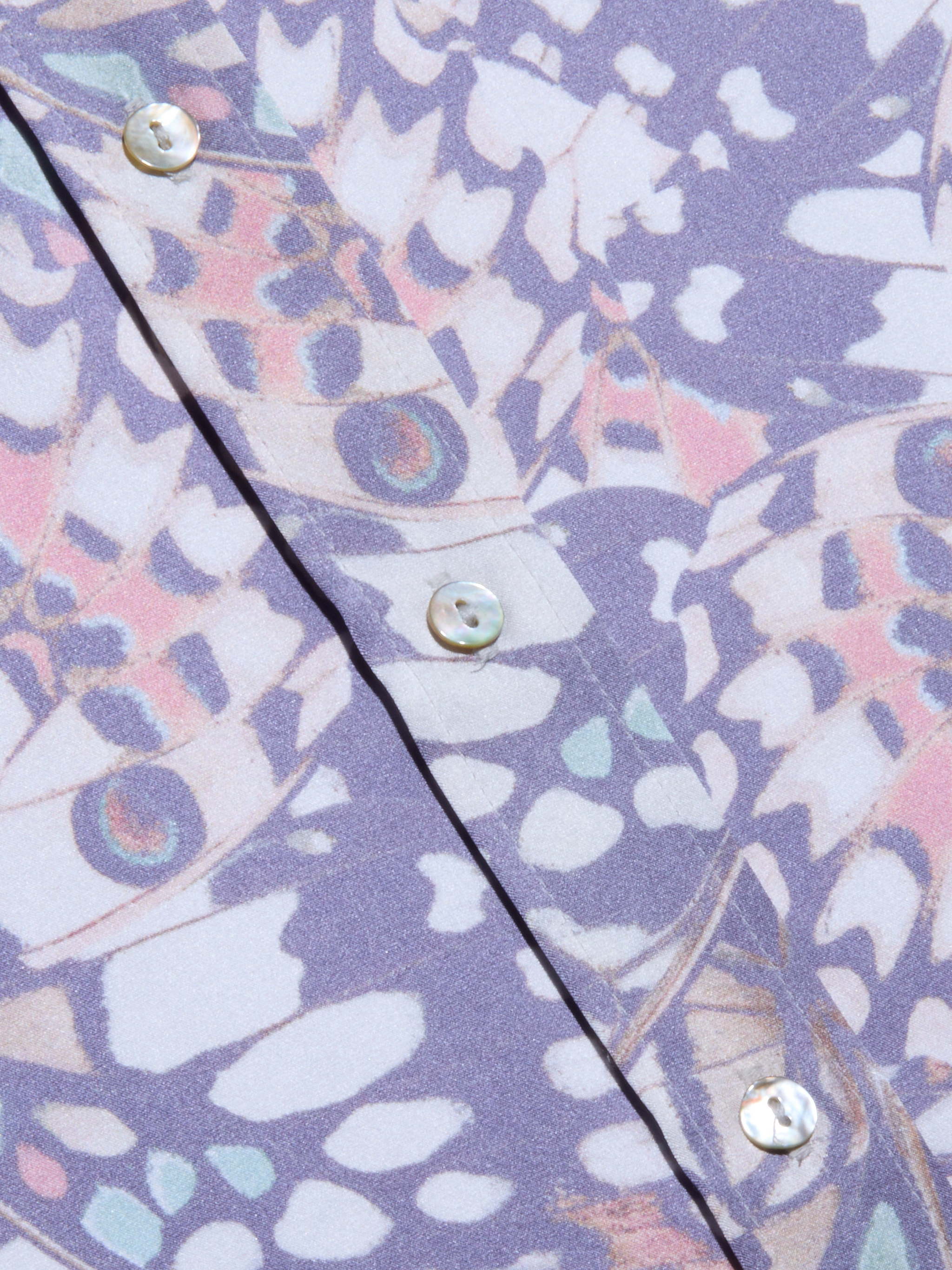 Primo piano del tessuto della blusa Dani Silk con motivi astratti ispirati alle ali delle farfalle in viola, rosa e bianco, bottoni iridescenti lungo la cucitura e una morbida finitura in seta per un look organico e ispirato alla natura.