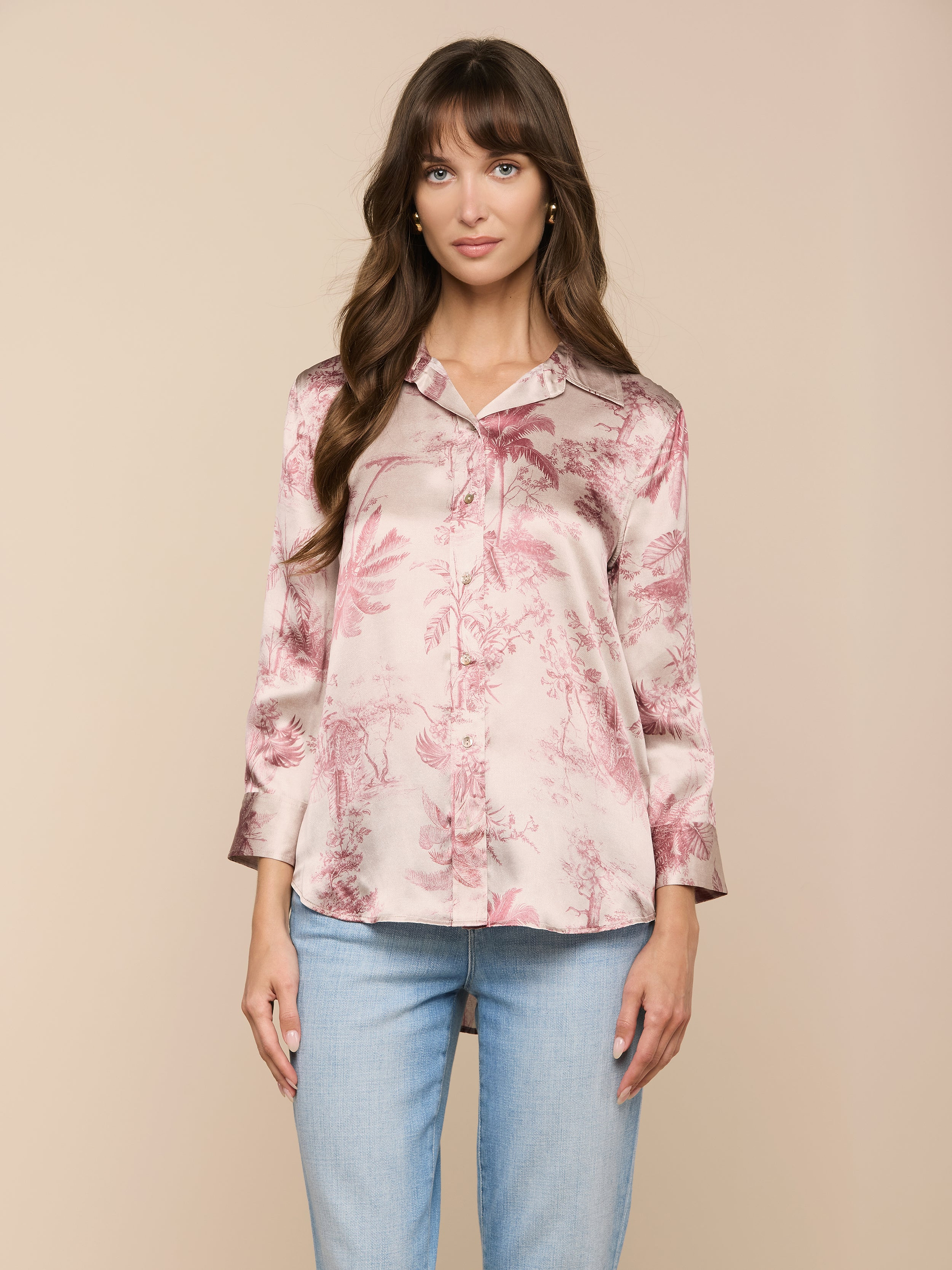 Eine Frau mit langen braunen Haaren trägt eine rosafarbene Dani-Seidenbluse mit einem dezenten Dschungel-Toile-Print und hellblaue Jeans. Sie steht vor einem beigen Hintergrund und blickt mit einem neutralen Gesichtsausdruck nach vorne.