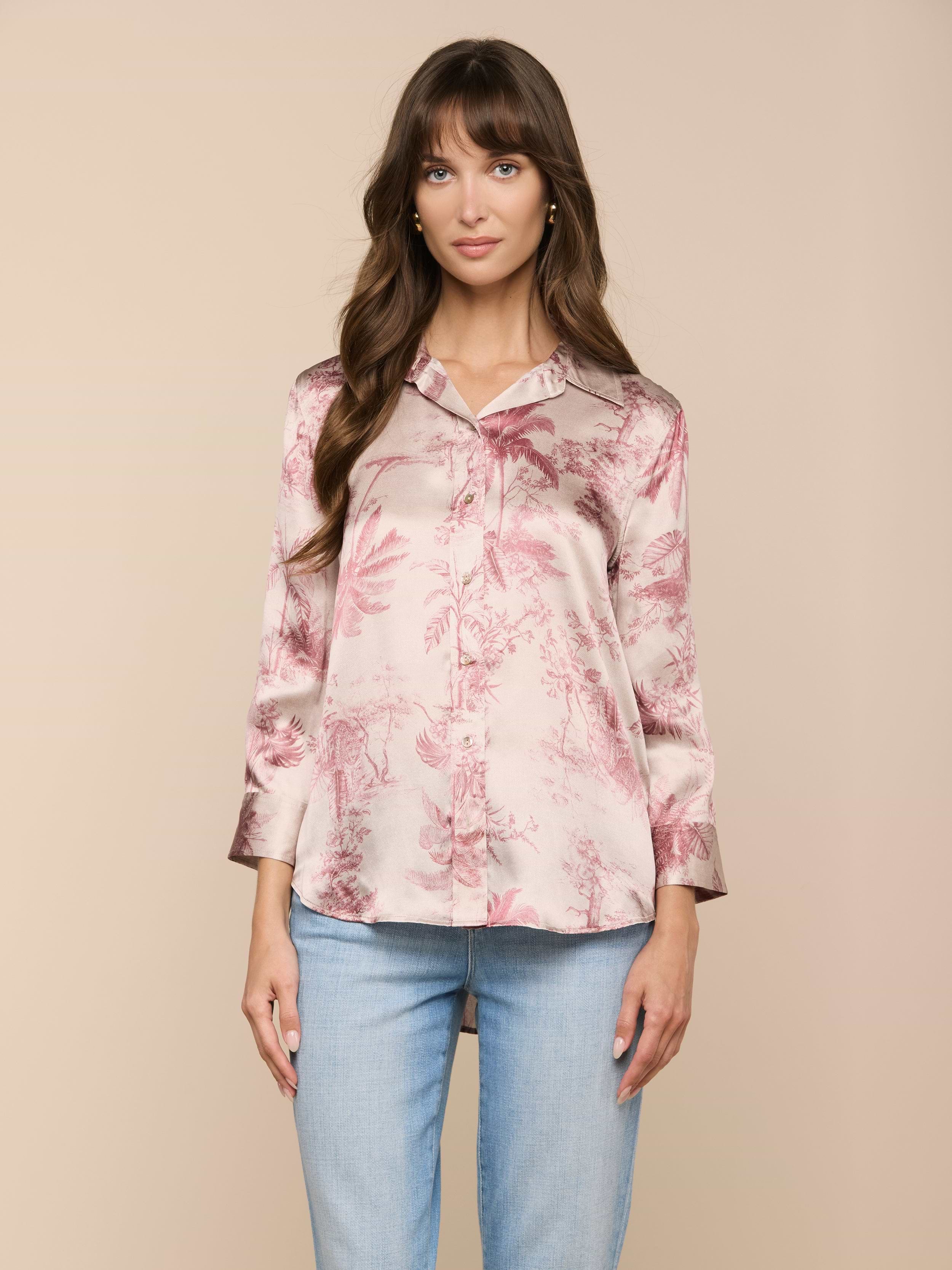 Eine Frau mit langen braunen Haaren trägt eine rosafarbene Dani-Seidenbluse mit einem dezenten Dschungel-Toile-Print und hellblaue Jeans. Sie steht vor einem beigen Hintergrund und blickt mit einem neutralen Gesichtsausdruck nach vorne.