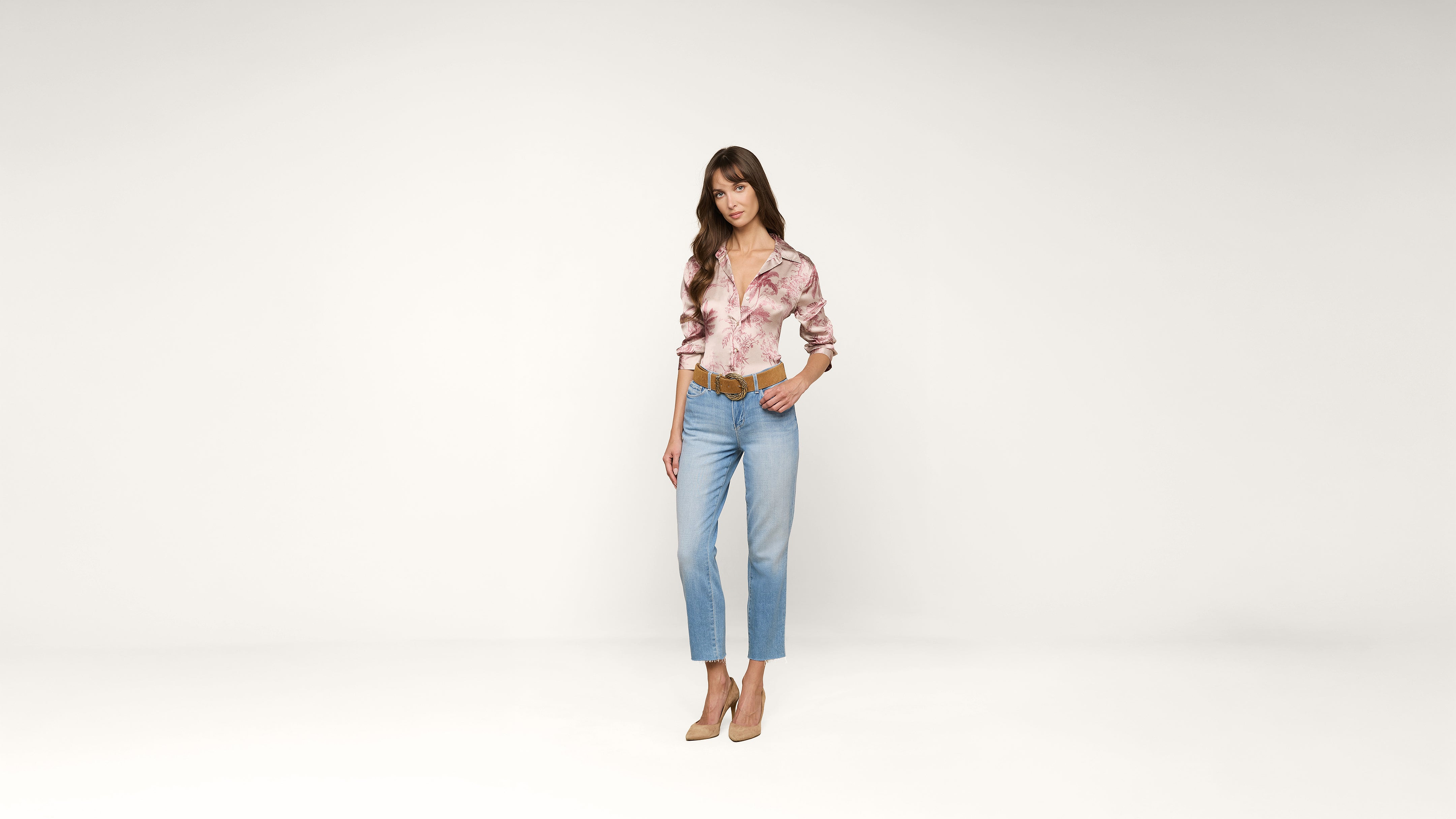 A woman stands in a minimal studio, wearing a pink patterned blouse, light blue jeans, tan belt, and tan heels. She poses with one hand in her pocket against a white background.