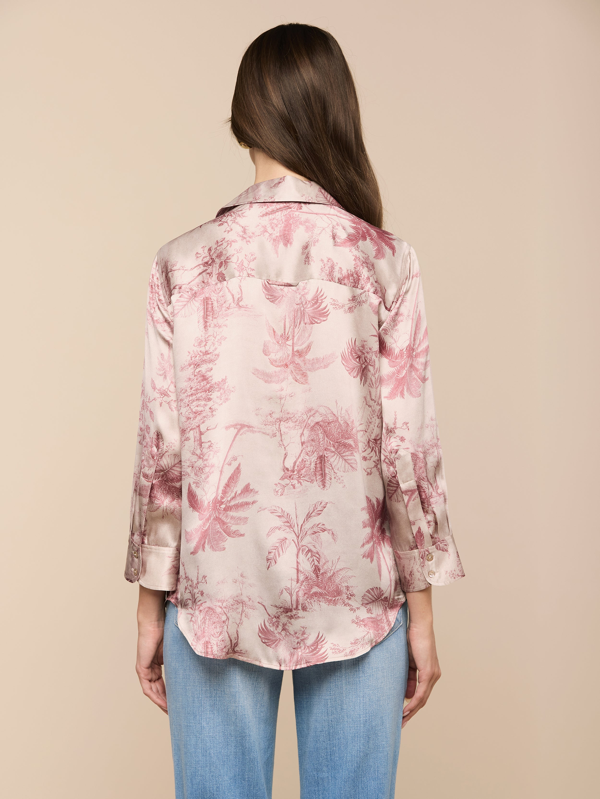 Eine Frau mit langen braunen Haaren, von hinten gesehen, trägt eine Dani-Seidenbluse mit Dschungel-Toile-Print und hellblaue Jeans vor einem beigen Hintergrund.