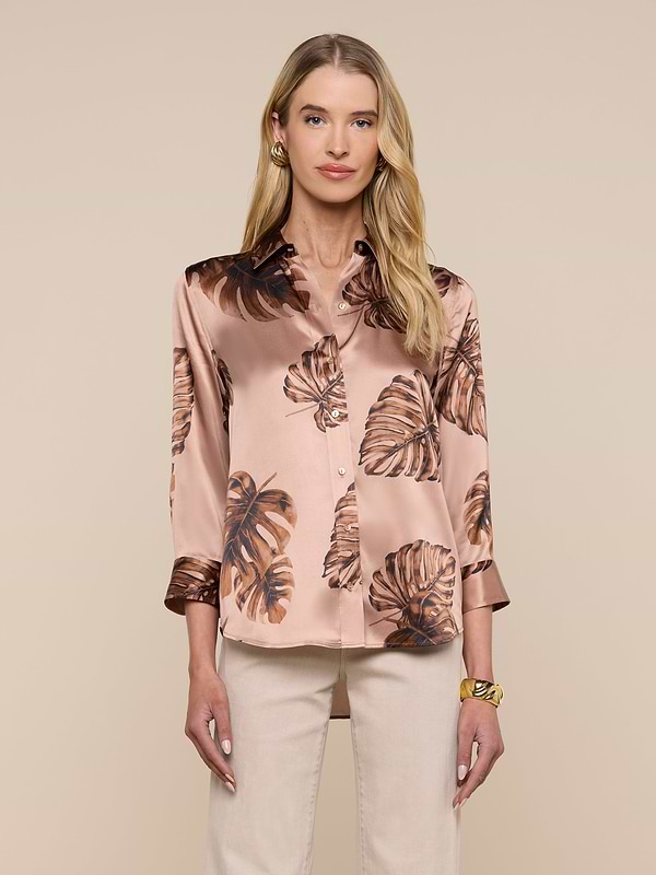 A woman stands facing forward against a plain light background, wearing the Dani Silk Blouse—a light pink, silky top adorned with a brown palm leaf print. She has long blonde hair and pairs the blouse with light-colored pants and a gold bracelet.