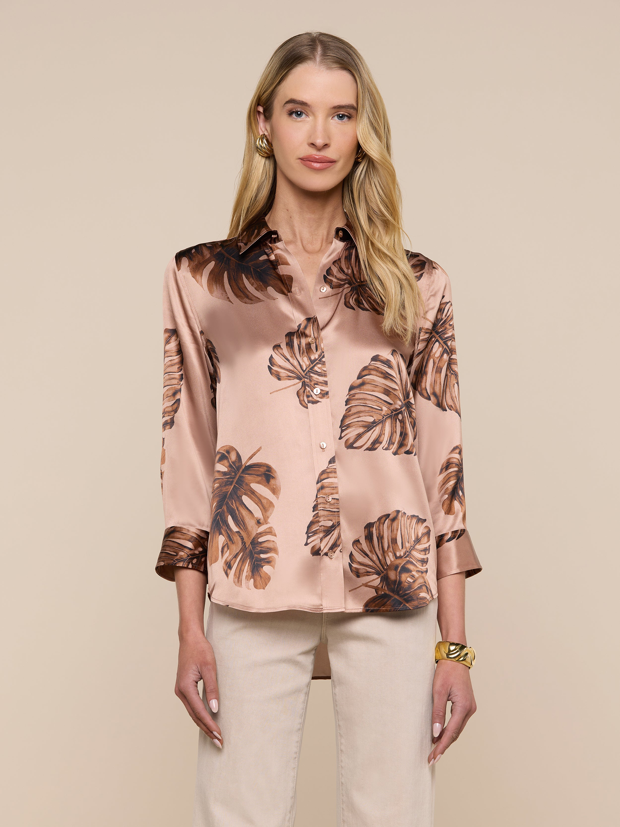 A woman stands facing forward against a plain light background, wearing the Dani Silk Blouse—a light pink, silky top adorned with a brown palm leaf print. She has long blonde hair and pairs the blouse with light-colored pants and a gold bracelet.