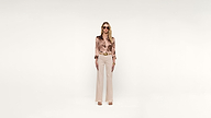 A woman stands facing forward, wearing sunglasses, a silky floral blouse, beige high-waisted pants with a belt, heeled sandals, and jewelry, all set against a plain white background.