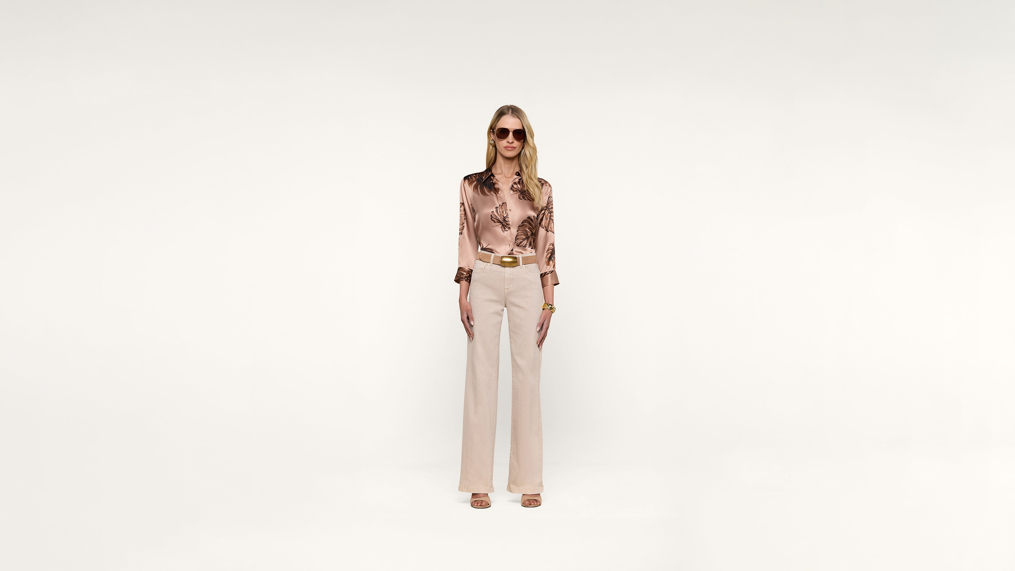 A woman stands facing forward, wearing sunglasses, a silky floral blouse, beige high-waisted pants with a belt, heeled sandals, and jewelry, all set against a plain white background.