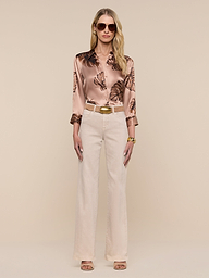 A woman with long blonde hair wears sunglasses, the Dani Silk Blouse featuring a palm leaf print, beige high-waisted pants, and nude heels, standing against a plain white background.