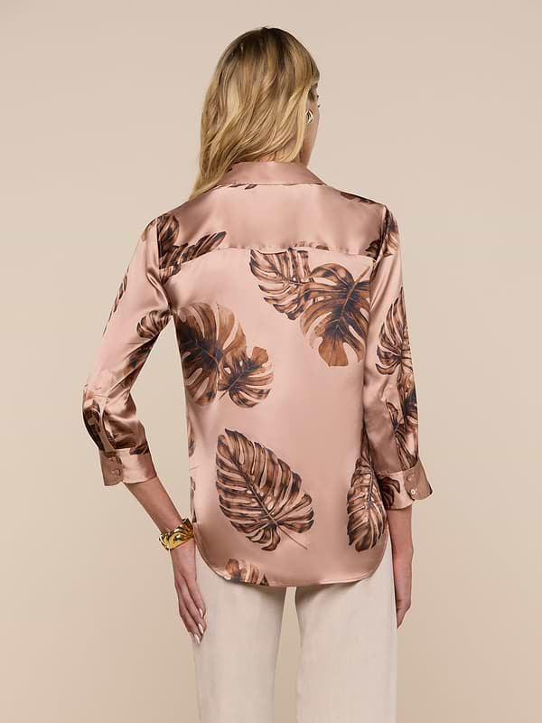 A woman with long blonde hair is seen from the back, wearing a shiny Dani Silk Blouse in light brown with a dark brown palm leaf print, paired with light beige pants, set against a plain light background.