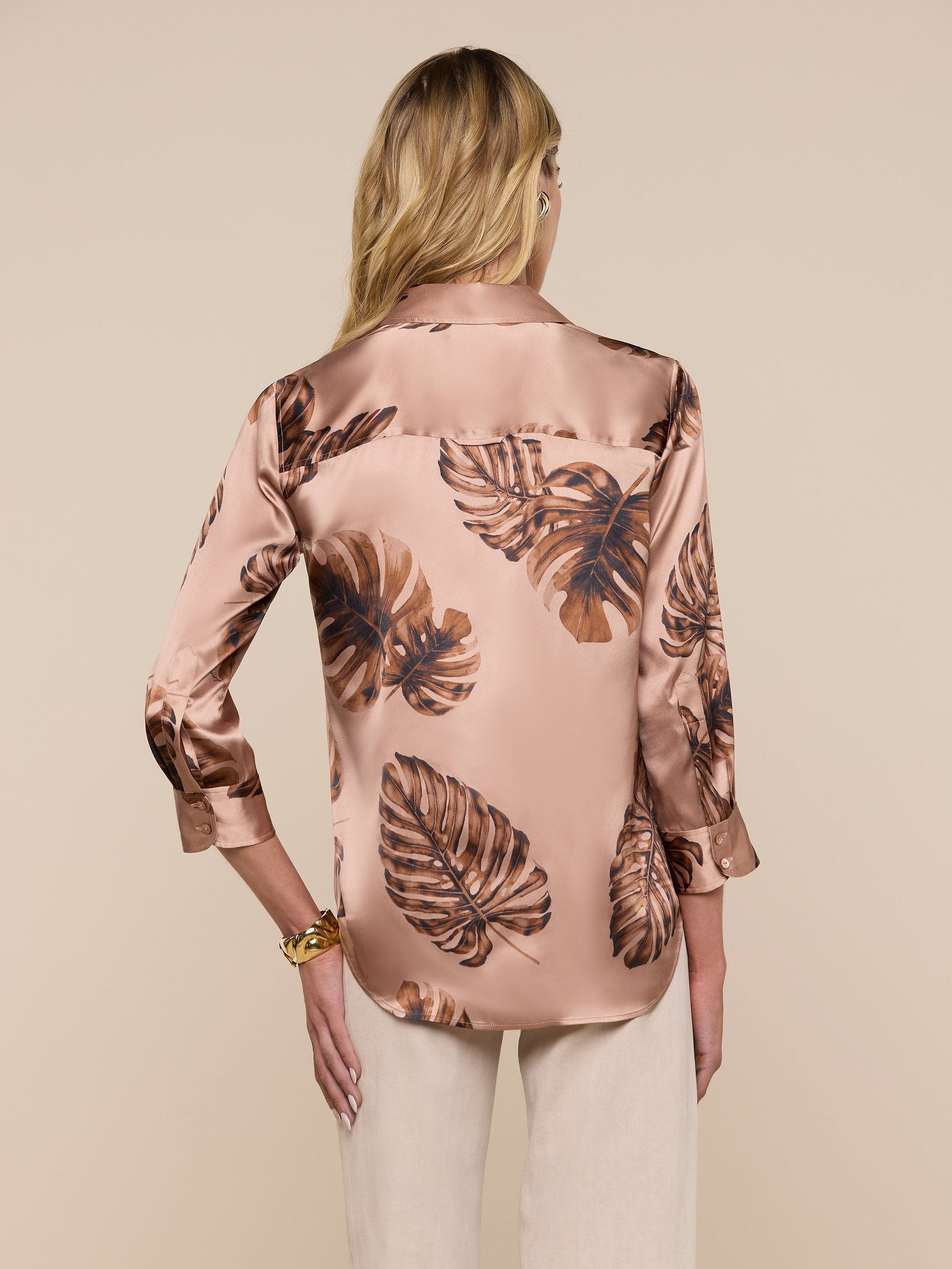 A woman with long blonde hair is seen from the back, wearing a shiny Dani Silk Blouse in light brown with a dark brown palm leaf print, paired with light beige pants, set against a plain light background.