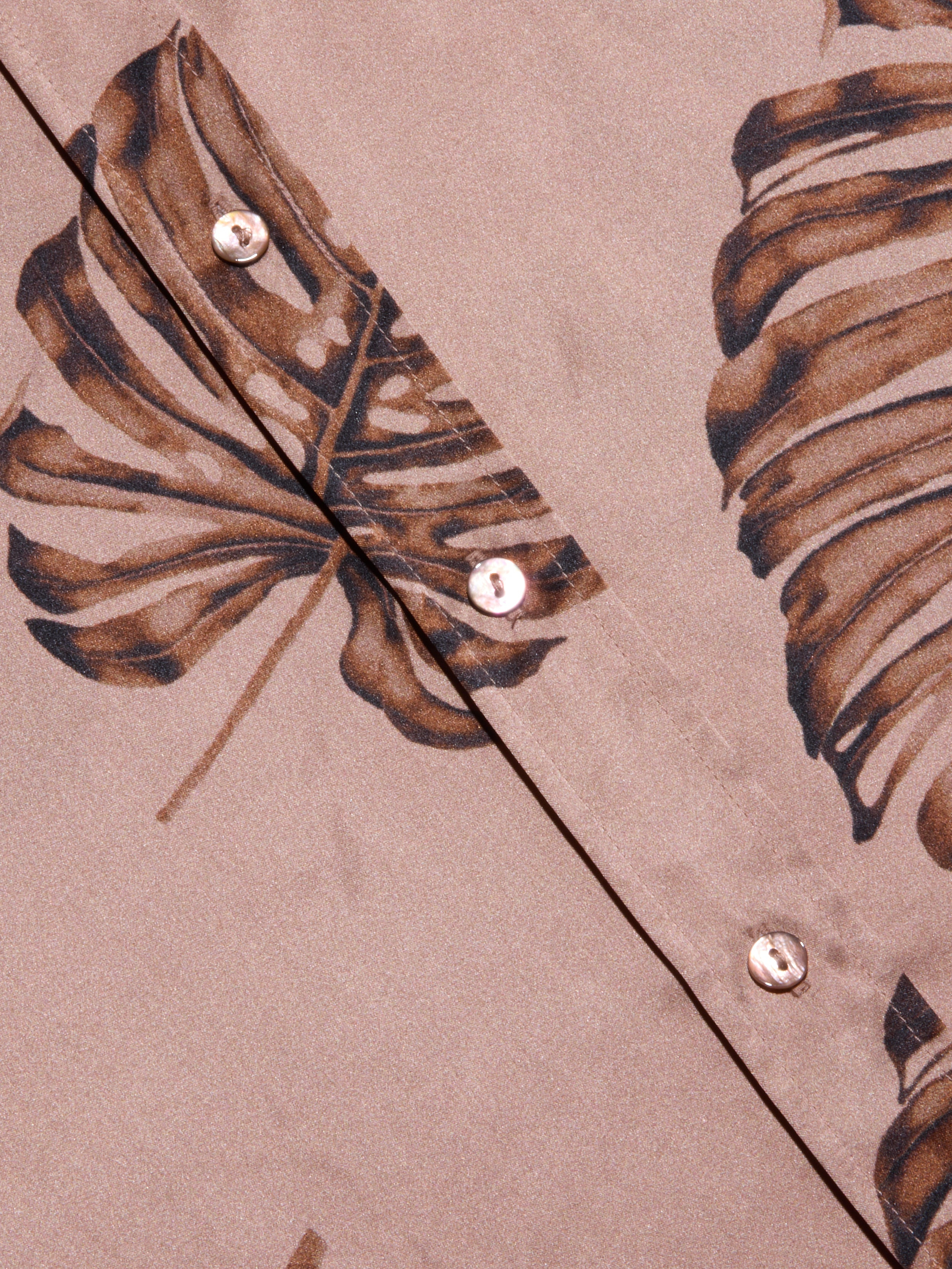 Close-up of the Dani Silk Blouse in beige, featuring a brown palm leaf print and pearl-like buttons. The smooth silk fabric and bold, tropical-inspired design give this button-down a distinctive look.