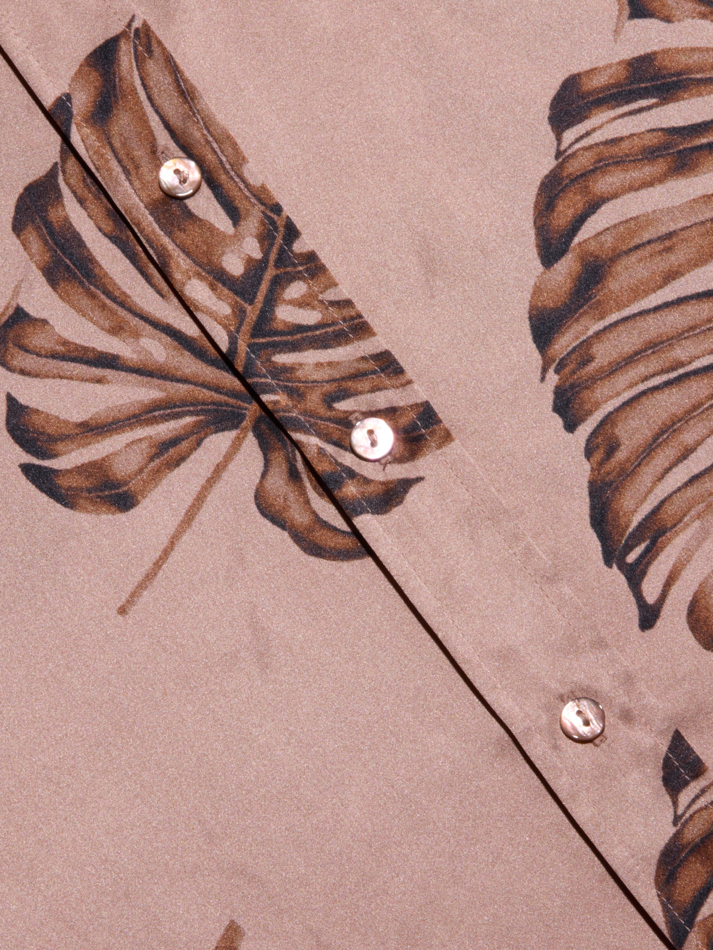 Close-up of the Dani Silk Blouse in beige, featuring a brown palm leaf print and pearl-like buttons. The smooth silk fabric and bold, tropical-inspired design give this button-down a distinctive look.