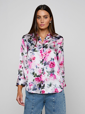 A woman with long brown hair wears the Dani Silk Blouse, a shiny button-down featuring a pink rose print and purple floral details, paired with blue jeans. She stands against a plain light gray background.