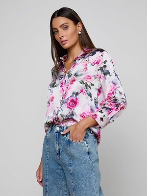 A woman stands against a plain background wearing the Dani Silk Blouse in pink rose print, paired with light blue high-waisted jeans. She has one hand in her pocket and a neutral expression.