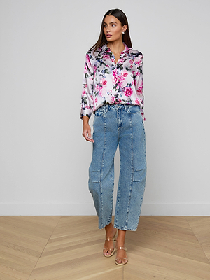 A woman in a pink rose print Dani Silk Blouse with long sleeves, light blue high-waisted wide-leg jeans, and clear heeled sandals stands against a plain white wall on a wooden floor.