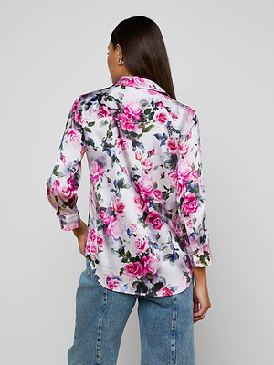 A woman with long brown hair wears the Dani Silk Blouse in a pink rose print, paired with blue jeans. She stands facing away from the camera against a plain background.
