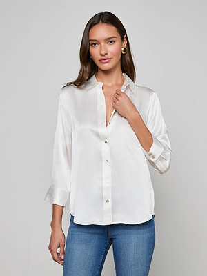 A woman with long brown hair wears the L'AGENCE Dani Silk Blouse in ivory, styled with rolled-up sleeves and blue jeans. She stands against a plain light gray background, looking at the camera with a slight smile.