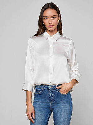 A woman with long brown hair wears the L'AGENCE Dani Silk Blouse in ivory, sleeves rolled up, paired with blue jeans, standing against a plain light gray background with a neutral expression.