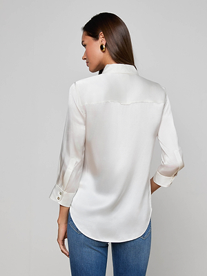 A woman with long brown hair is seen from behind wearing the L'AGENCE Dani Silk Blouse in ivory with rolled-up sleeves and blue jeans, standing against a plain light gray background.