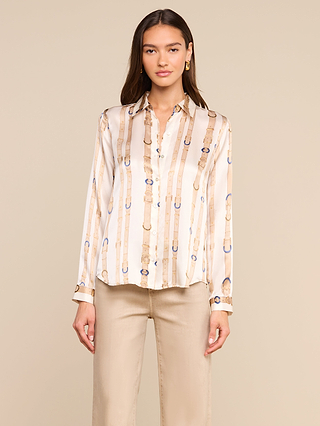 A woman stands against a beige background, wearing the cream Tyler Silk Blouse by L’AGENCE featuring a vertical belt pattern, paired with beige pants. She has straight brown hair and gazes at the camera with a neutral expression.