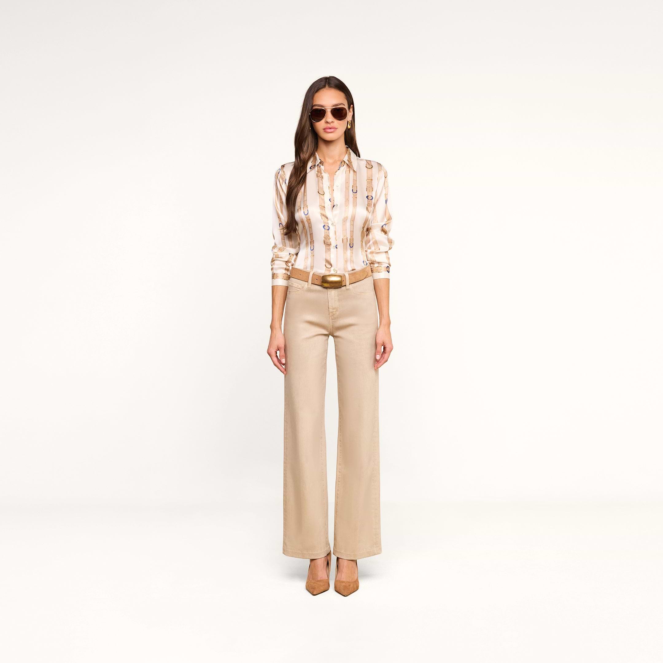 A woman wearing sunglasses, the Tyler Silk Blouse from L’AGENCE, beige wide-leg pants, and heels stands facing forward in a minimal white studio.