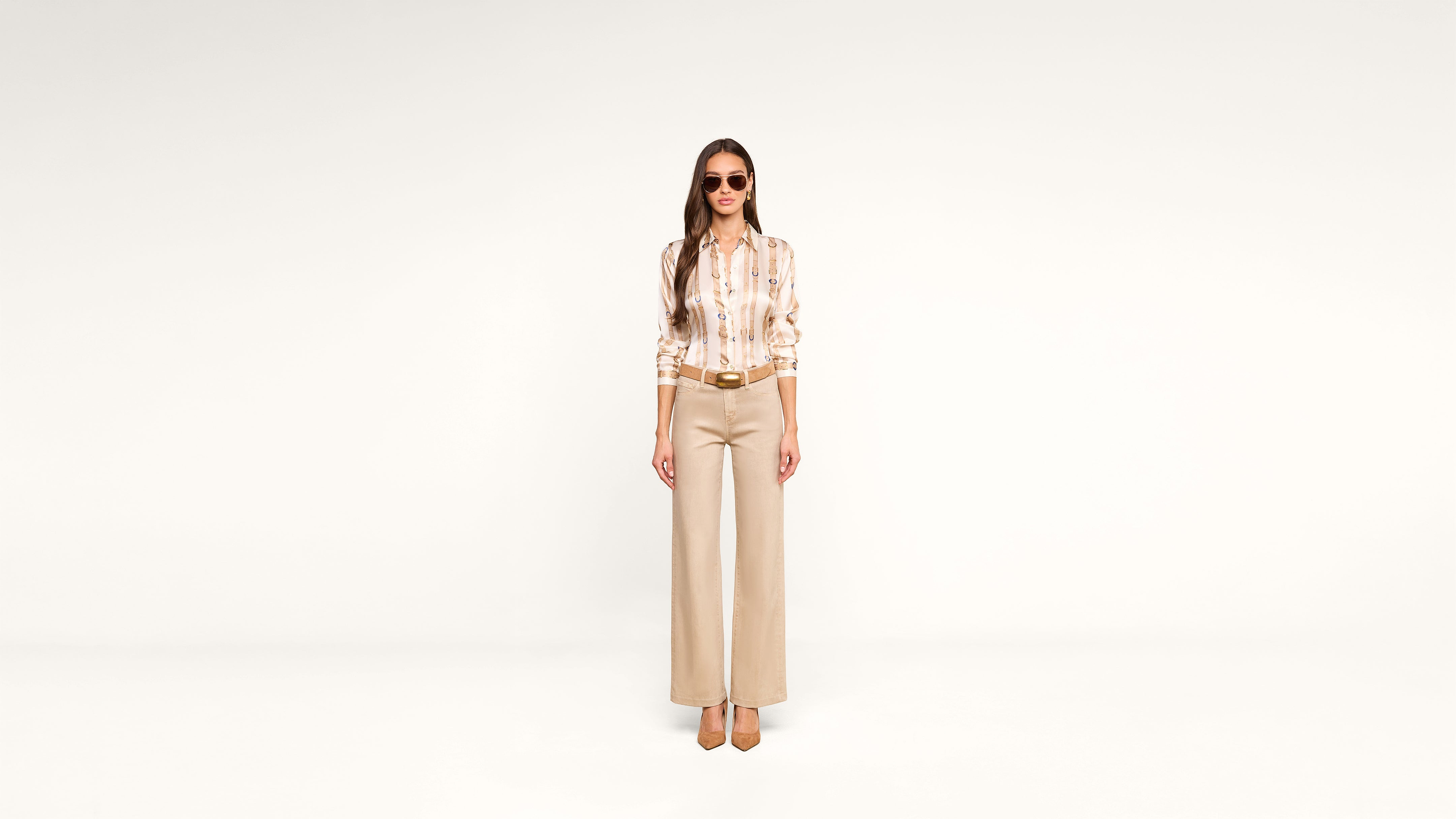 Woman wearing sunglasses, a patterned blouse, beige wide-leg pants, and heels stands facing forward against a plain white background.