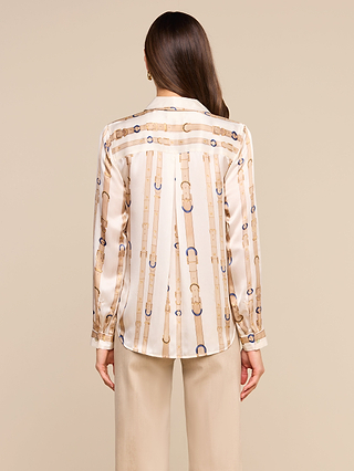 A woman stands with her back to the camera, wearing the Tyler Silk Blouse by LAGENCE, featuring a beige and blue chain print and silky long sleeves. She pairs it with light-colored pants against a plain beige background.