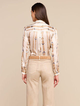 A woman stands with her back to the camera, wearing the Tyler Silk Blouse by LAGENCE—featuring a belt pattern and long sleeves—paired with light beige pants, set against a plain beige background.