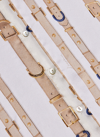 Patterned with rows of light brown belts, gold buckles, and small blue horseshoe accents on a white background, the Tyler Silk Blouse by L’AGENCE features visible fabric texture and button details that lend this long-sleeve shirt a refined yet playful touch.