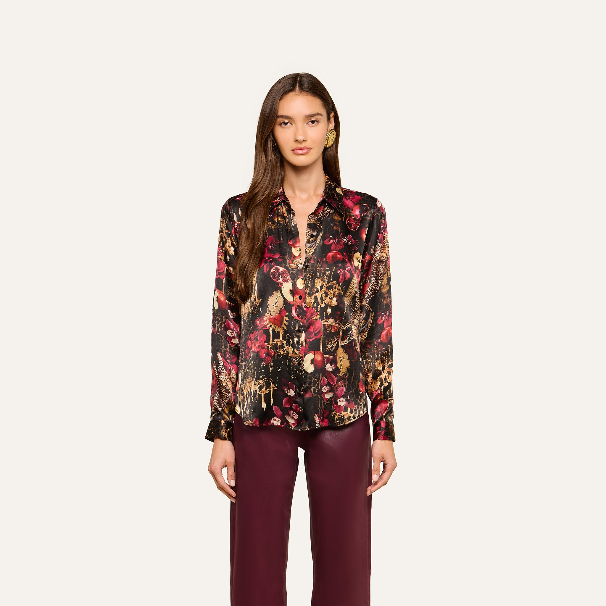 A woman wearing the Tyler Silk Blouse from L’AGENCE—a long-sleeve, button-up shirt featuring a colorful, abstract floral pattern in burgundy, gold, and black—paired with burgundy pants and standing against a plain light background.