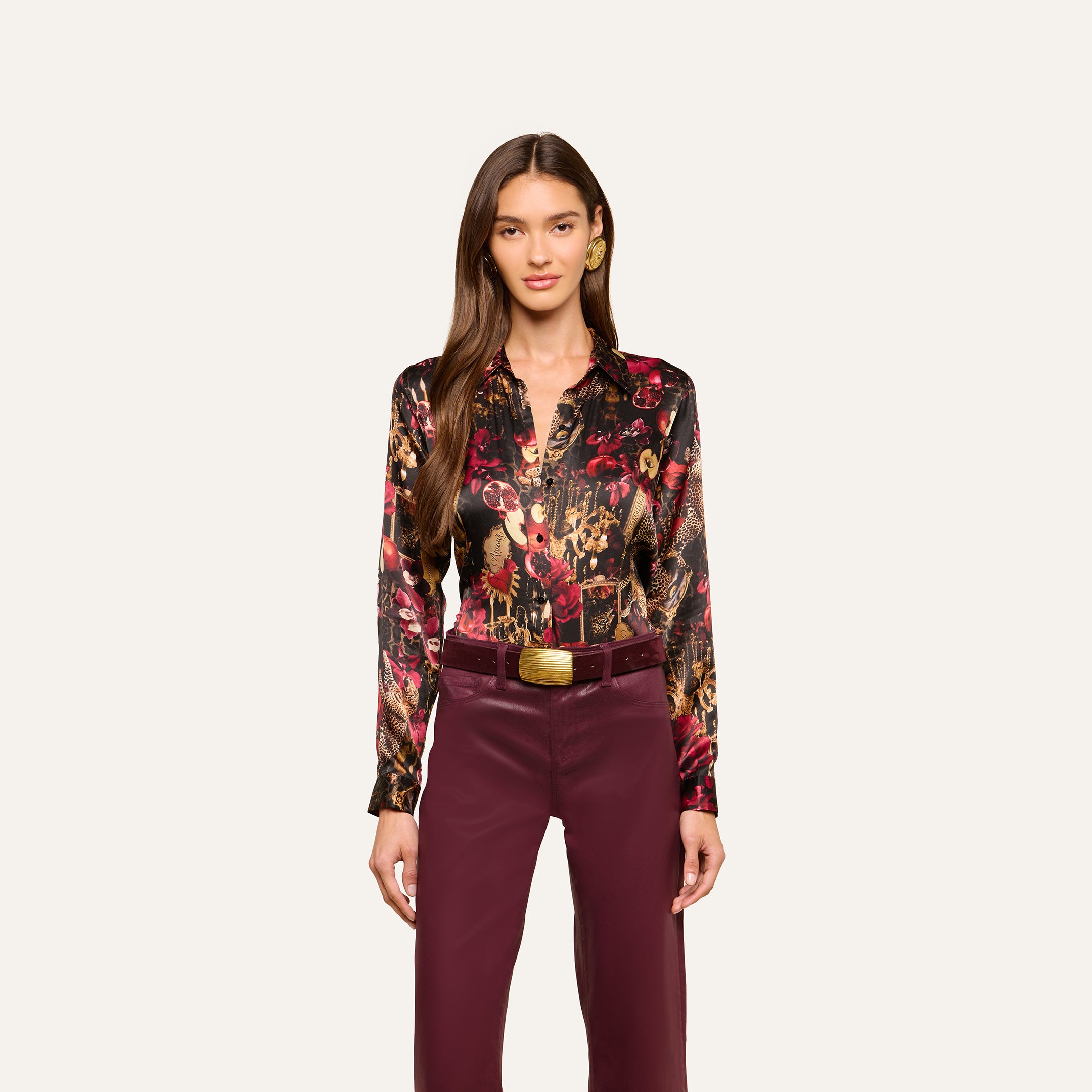 A woman stands facing forward against a plain light background, wearing the Tyler Silk Blouse from L’AGENCE—featuring long sleeves and a dark floral print—paired with burgundy pants and a wide belt with a gold buckle.