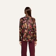 A woman stands facing away, wearing the Tyler Silk Blouse—a long-sleeve, multicolored floral top—paired with burgundy pants against a plain, light background.