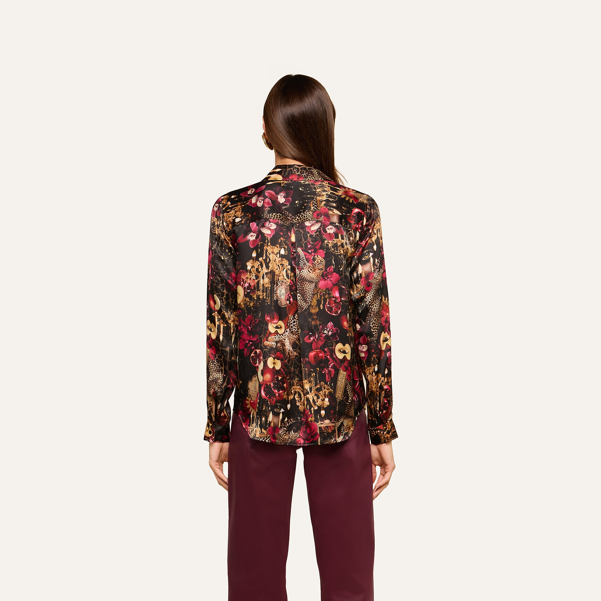 A woman stands facing away, wearing the Tyler Silk Blouse—a long-sleeve, multicolored floral top—paired with burgundy pants against a plain, light background.