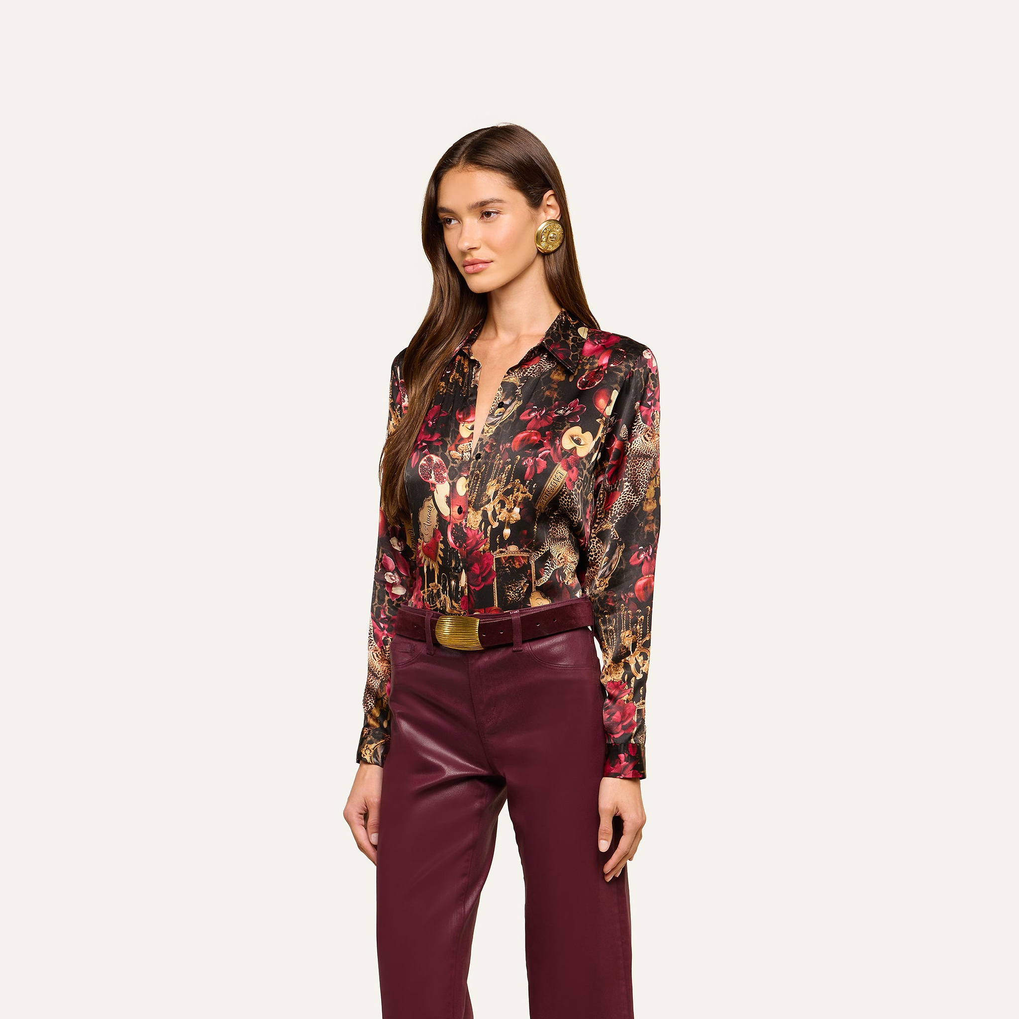 Wearing the Tyler Silk Blouse—a colorful, patterned L’AGENCE silk shirt with red and brown tones and long sleeves—the woman pairs it with high-waisted burgundy pants and a wide belt, standing against a plain light background.