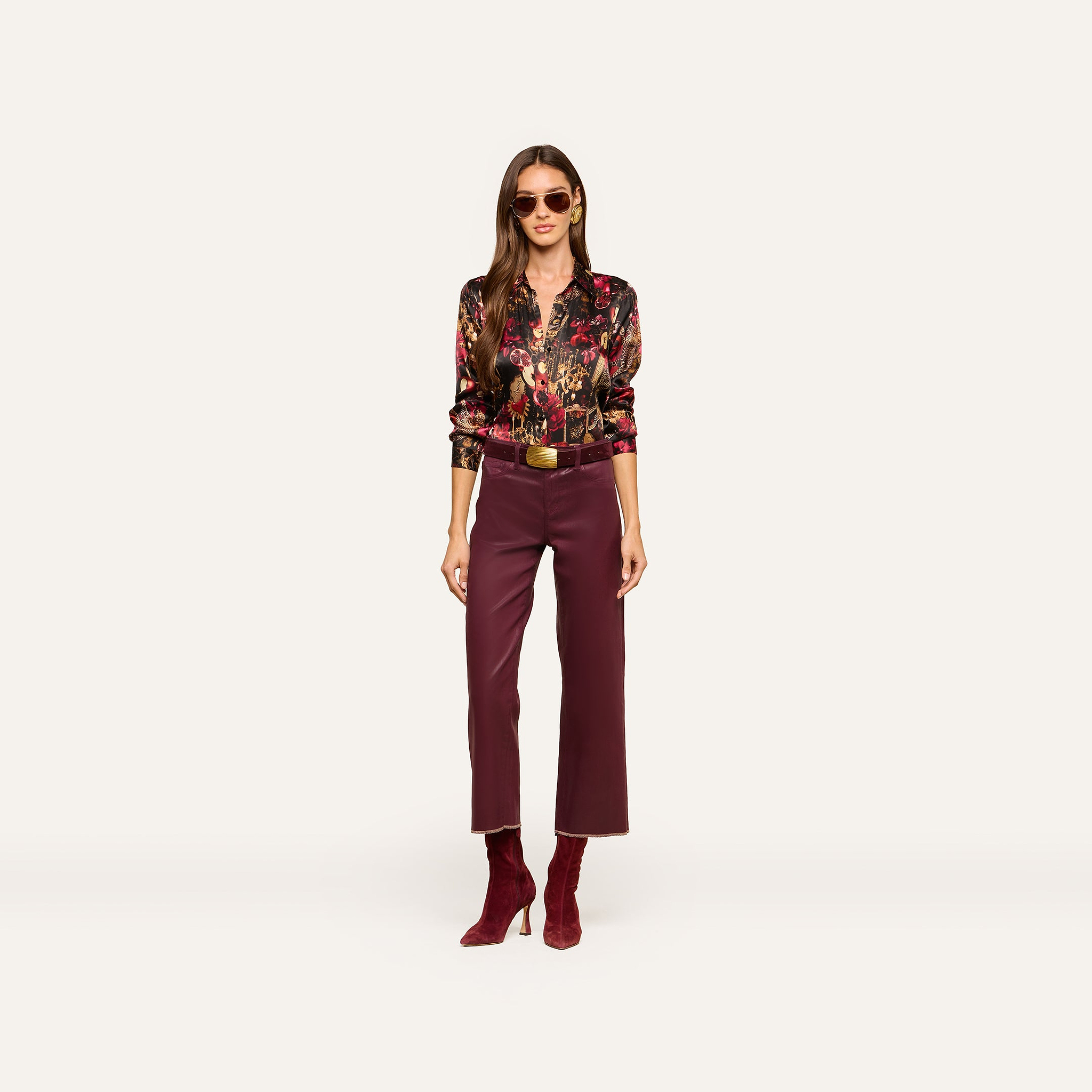 A woman wearing sunglasses, the Tyler Silk Blouse from L’AGENCE—a colorful floral silk top—maroon pants, and matching maroon boots stands facing forward against a plain light background.