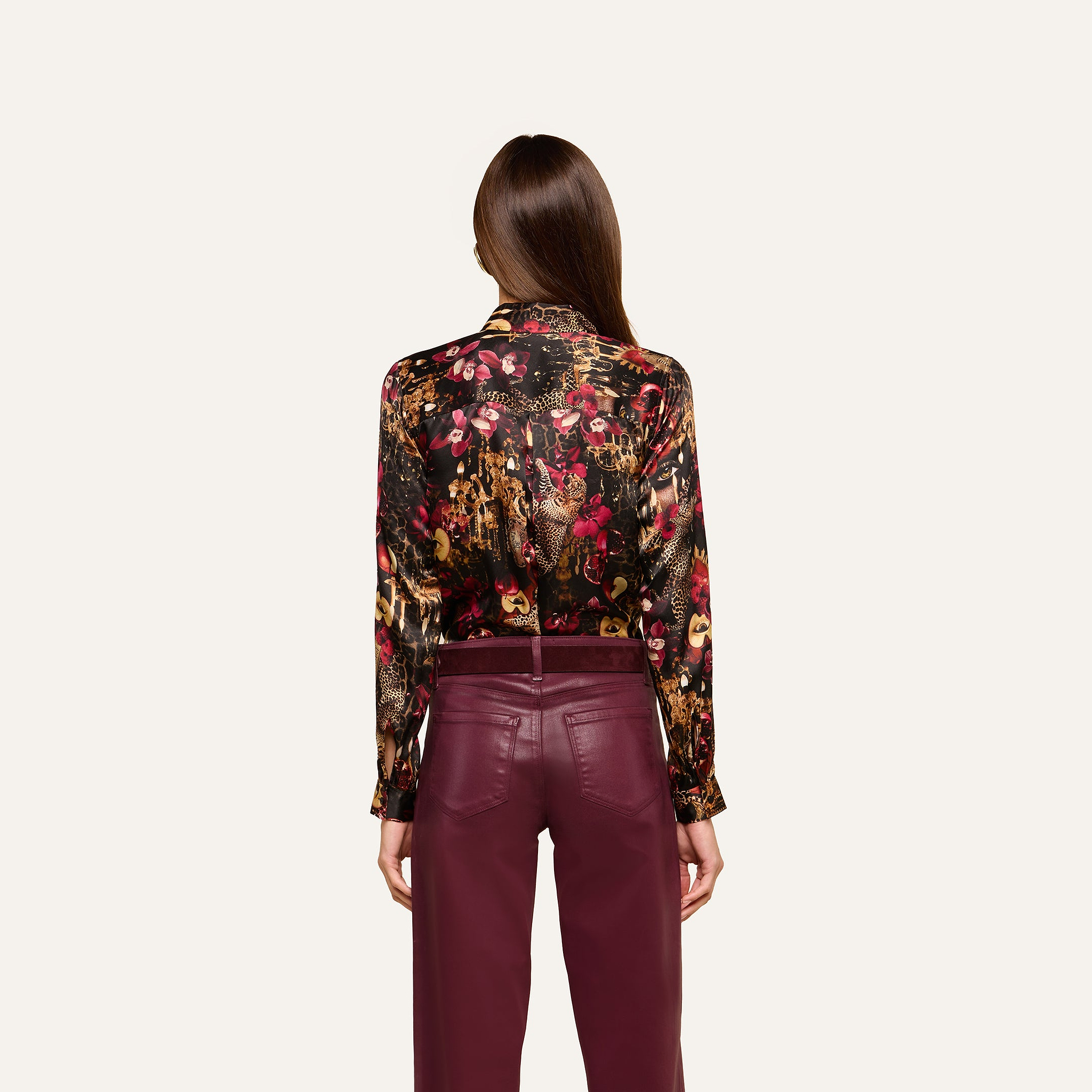 A woman with straight brown hair, worn down, stands against a plain light background, facing away and wearing the L’AGENCE Tyler Silk Blouse featuring a colorful floral pattern paired with burgundy pants.