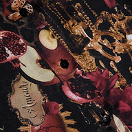 Close-up of the Tyler Silk Blouse from L’AGENCE, highlighting its luxurious patterned silk fabric adorned with pomegranates, apples, gold chains, and ornate decorative details. The design features black buttons sewn on top and includes a scroll labeled “Amour” amid the rich motifs.
