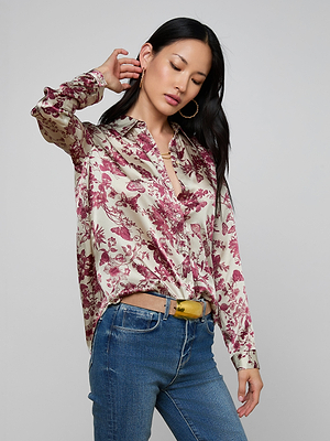 A woman with long dark hair and hoop earrings poses with one hand on her head, wearing a Tyler Silk Blouse featuring a red floral butterfly print, blue jeans, and a yellow belt against a plain light background.