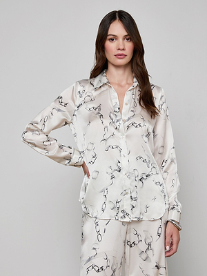 A woman with long, wavy brown hair and a neutral expression stands against a plain background, wearing the Tyler Silk Blouse in cream with a gray chain print and matching pants. One hand rests on her hip.