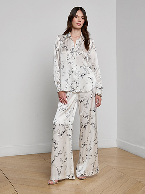 A woman with long brown hair stands in a minimalist room wearing a L'AGENCE Tyler Silk Blouse with a gray chain print, styled pajama-style with wide-leg pants and sandals.