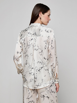 A woman faces away, head turned to the side, wearing the cream Tyler Silk Blouse with black abstract print and matching pants against a plain light gray background.