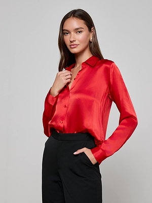 A woman with long brown hair models the L'AGENCE Tyler Silk Blouse in shiny red with black trousers, standing against a light gray background—a timeless staple for any woman’s wardrobe.
