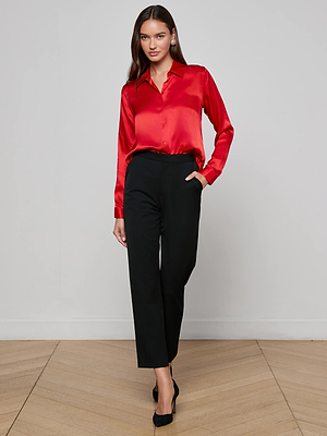 A woman with long brown hair stands against a plain wall wearing the L'AGENCE Tyler Silk Blouse in shiny red, paired with black cropped trousers and heels. One hand is in her pocket. This timeless blouse elevates any wardrobe.