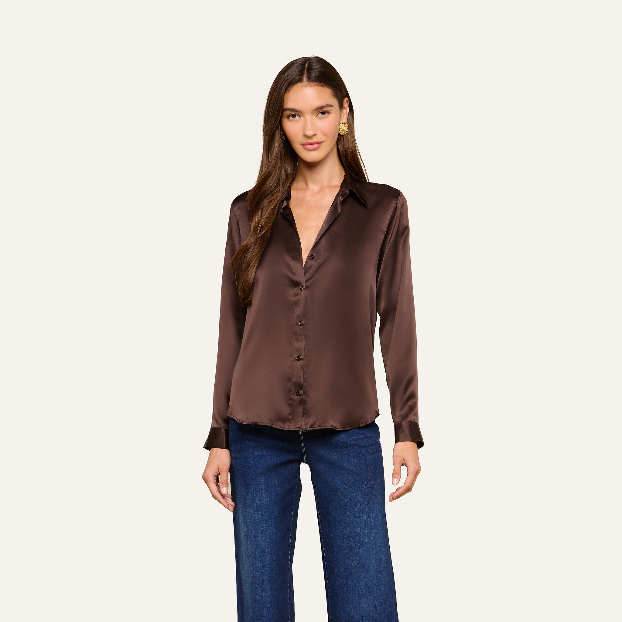 A woman wearing the Tyler Silk Blouse in dark brown, featuring long sleeves and a collar, paired with blue jeans and standing against a plain light background.