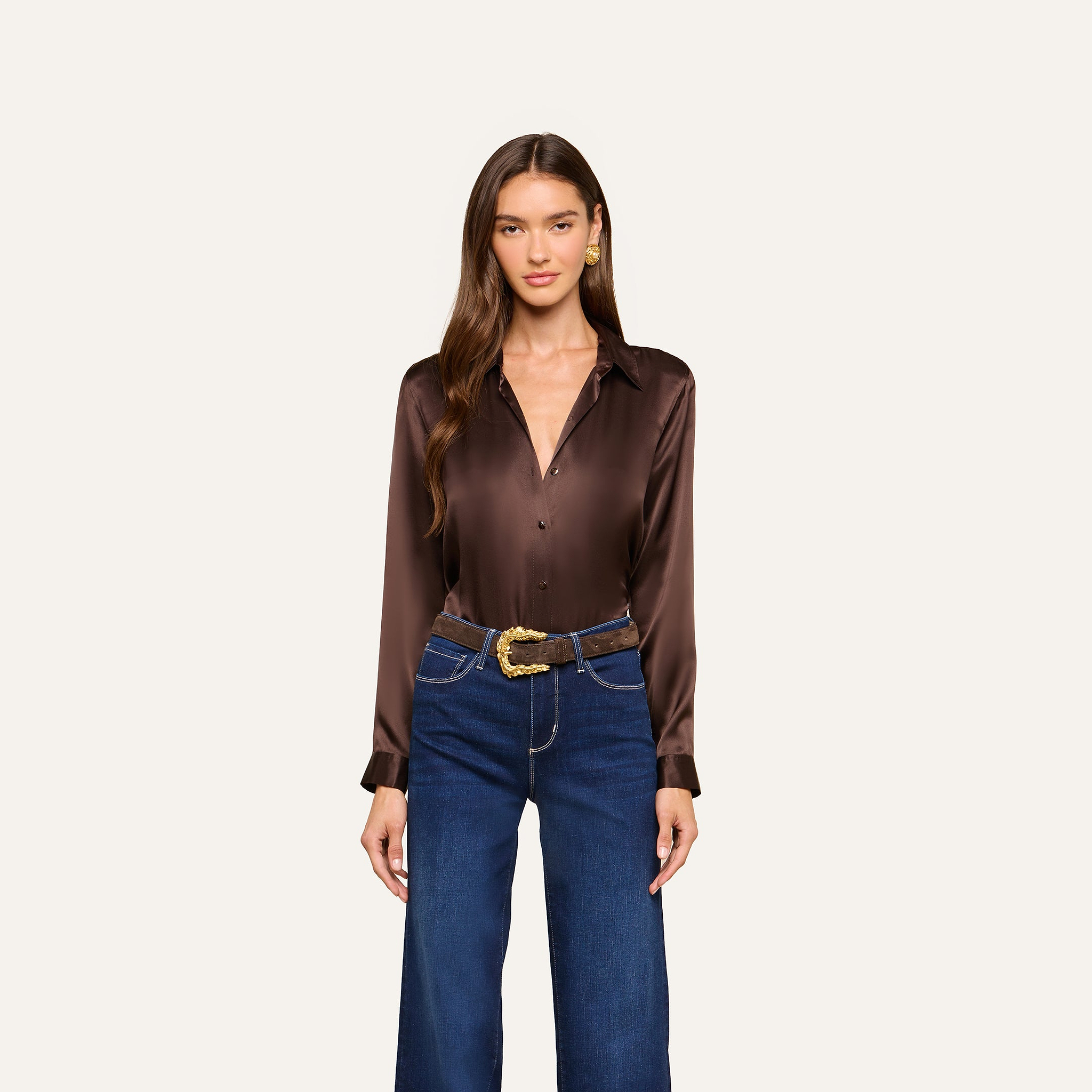 A woman with long brown hair wears the Tyler Silk Blouse in dark brown, paired with blue wide-leg jeans featuring white stitching and a decorative gold belt, standing against a plain light background.